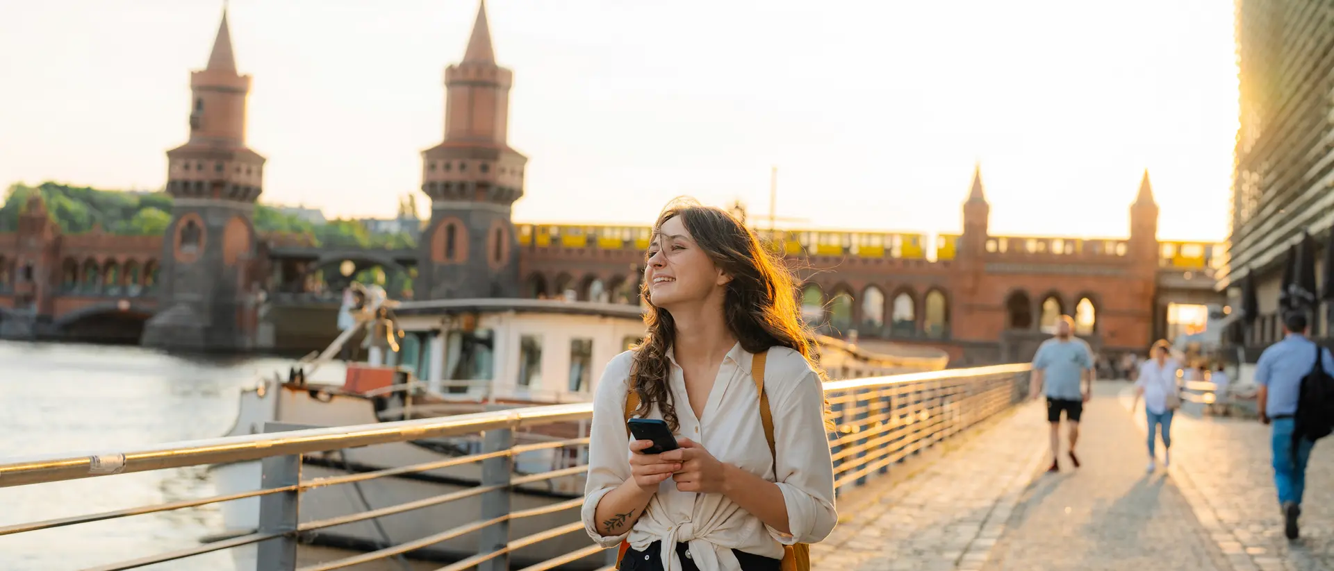 A woman stands on a riverside path, checking her phone, with a bridge and sunset in the background.