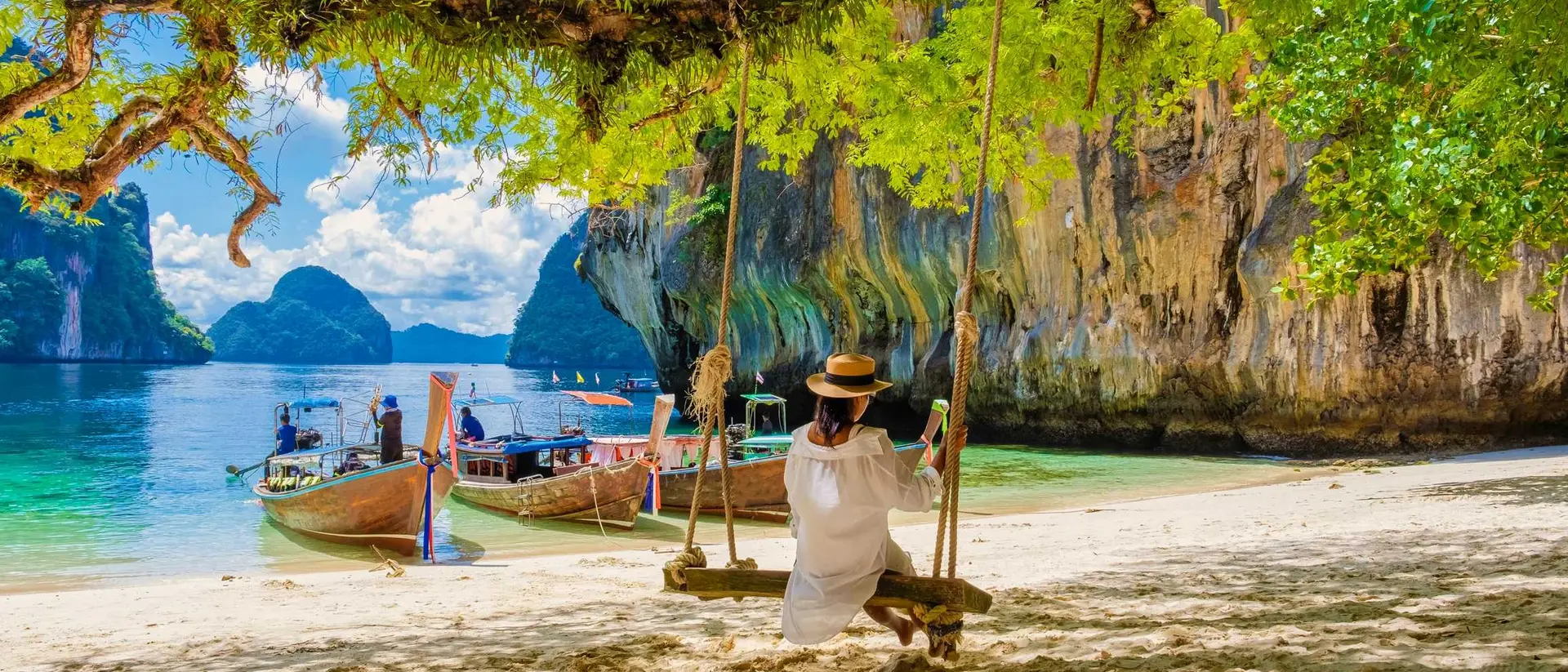 A person on a swing overlooking a tropical beach with boats and limestone cliffs in the background under a sunny sky.