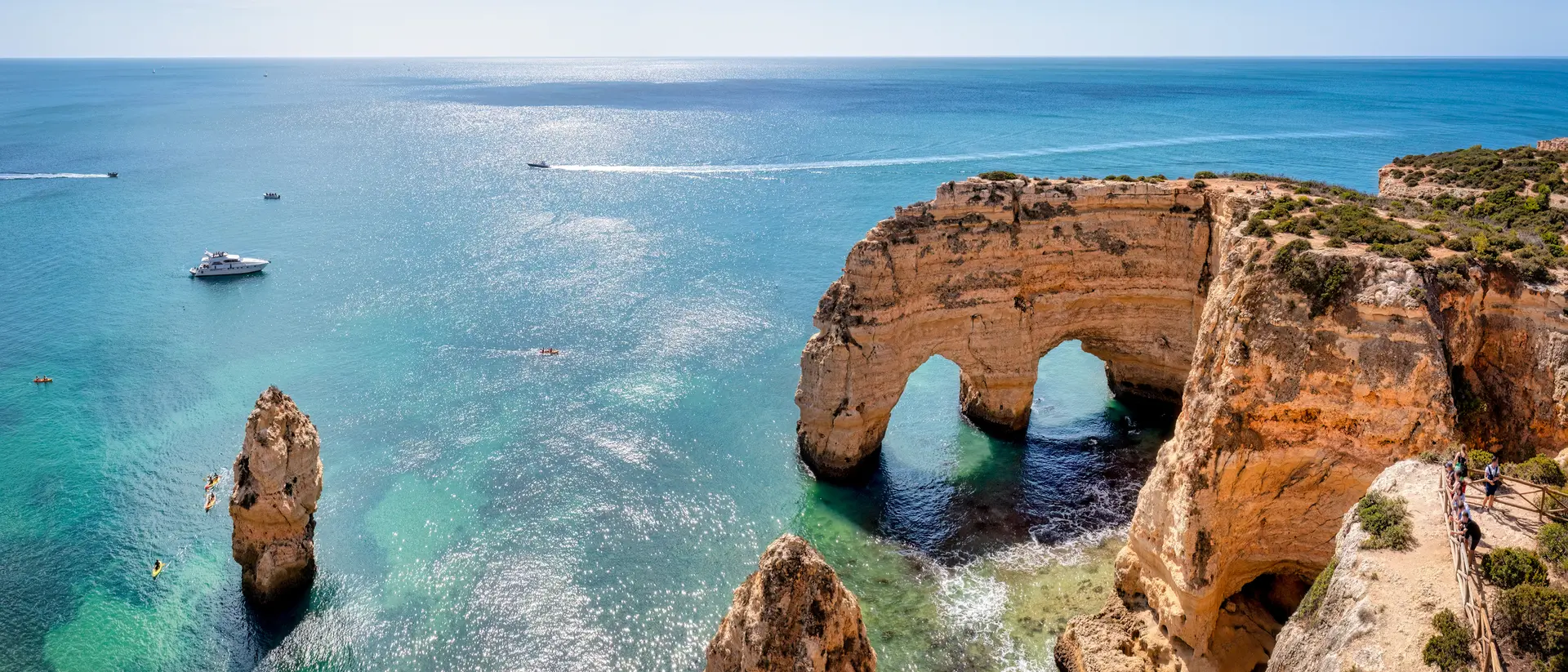 Scenic view of sea cliffs with arches and rock formations, boats on clear blue water under a sunny sky.