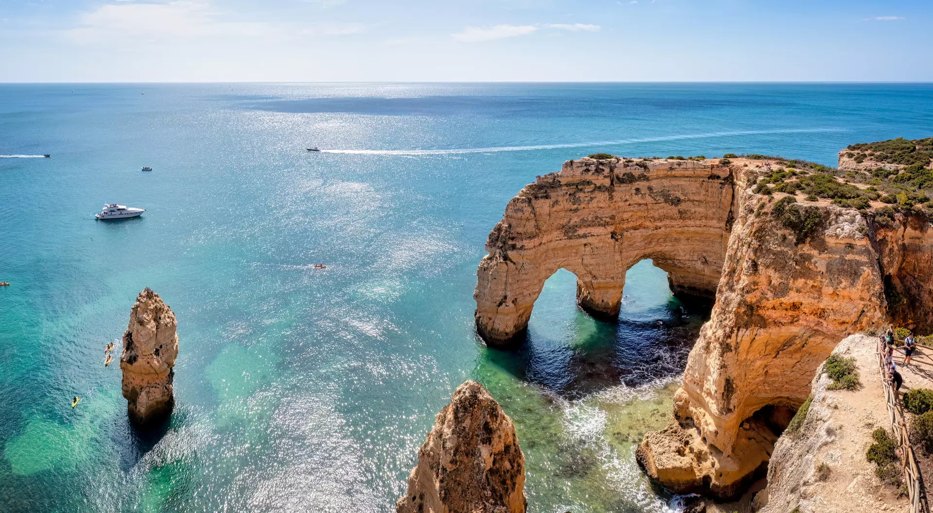 Scenic view of sea cliffs with arches and rock formations, boats on clear blue water under a sunny sky.