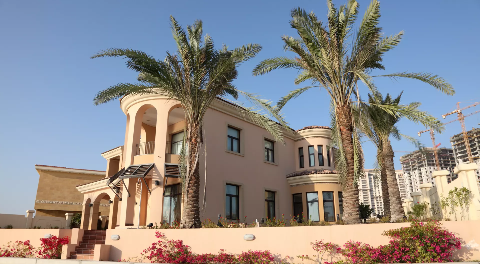 Peach-colored house with palm trees and pink flowers in the foreground, against a clear blue sky.