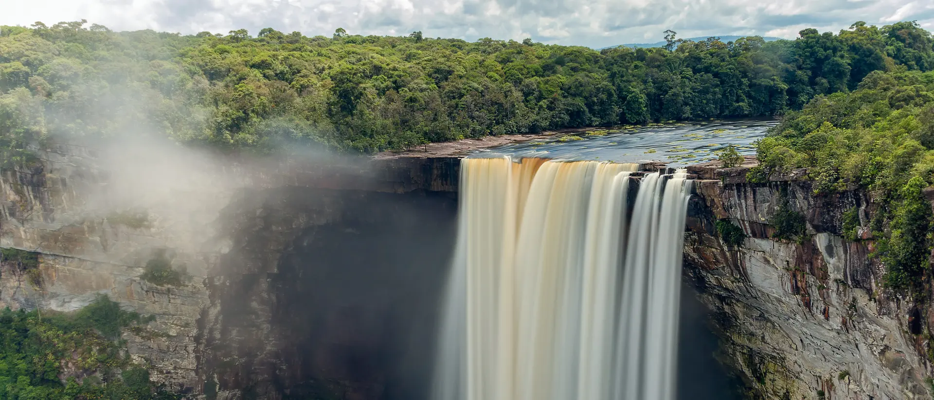 A waterfall cascades over a rocky cliff, surrounded by lush greenery and a blue sky with fluffy clouds.