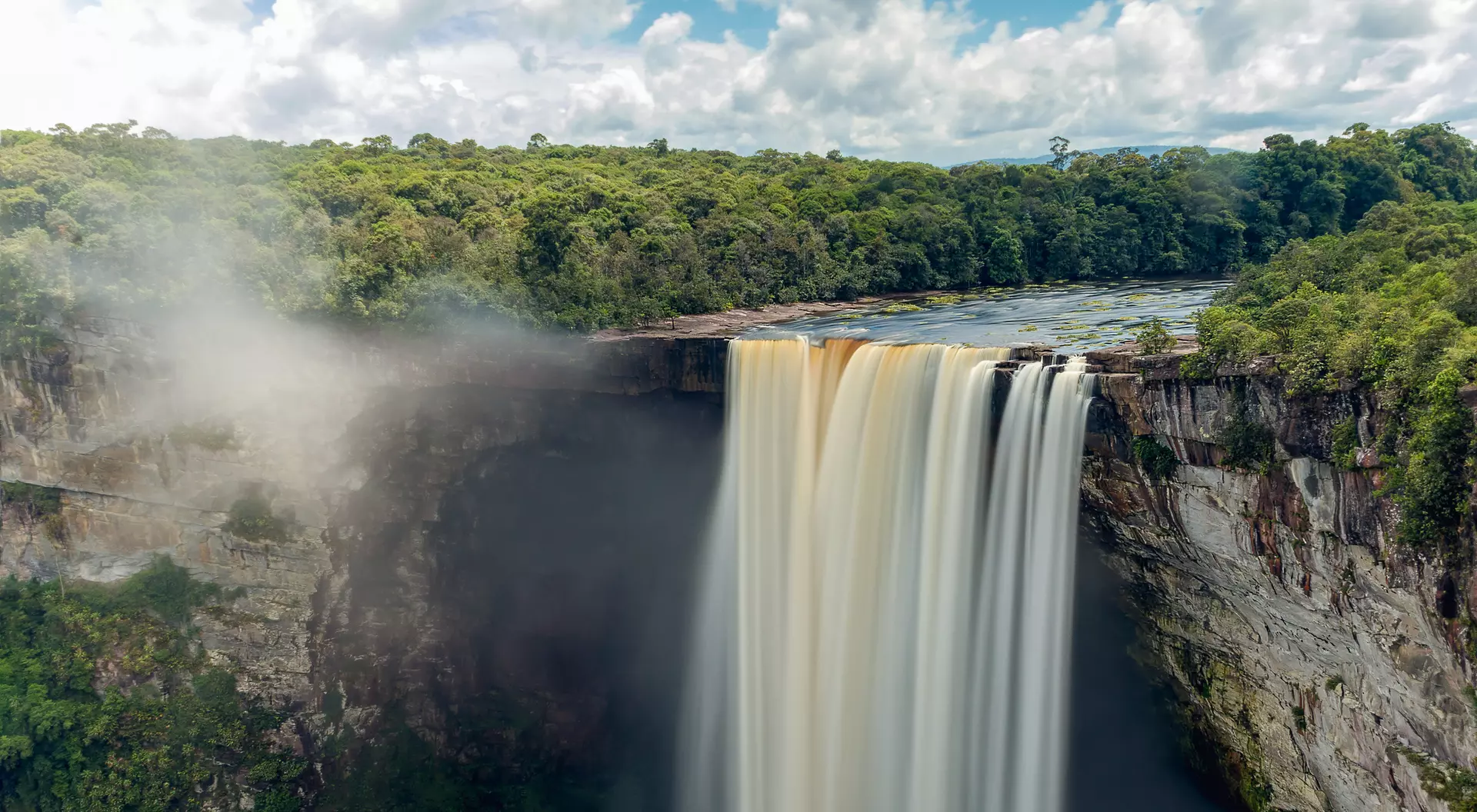 A waterfall cascades over a rocky cliff, surrounded by lush greenery and a blue sky with fluffy clouds.