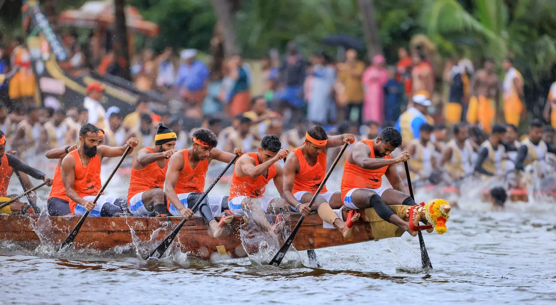 A team of rowers in orange shirts paddles a dragon boat, competing in a race with a large crowd watching.