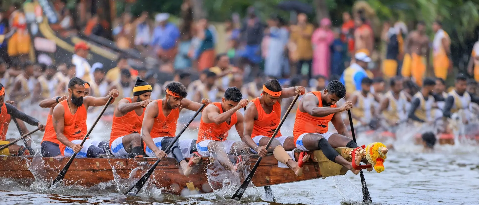 A team of rowers in orange shirts paddles a dragon boat, competing in a race with a large crowd watching.