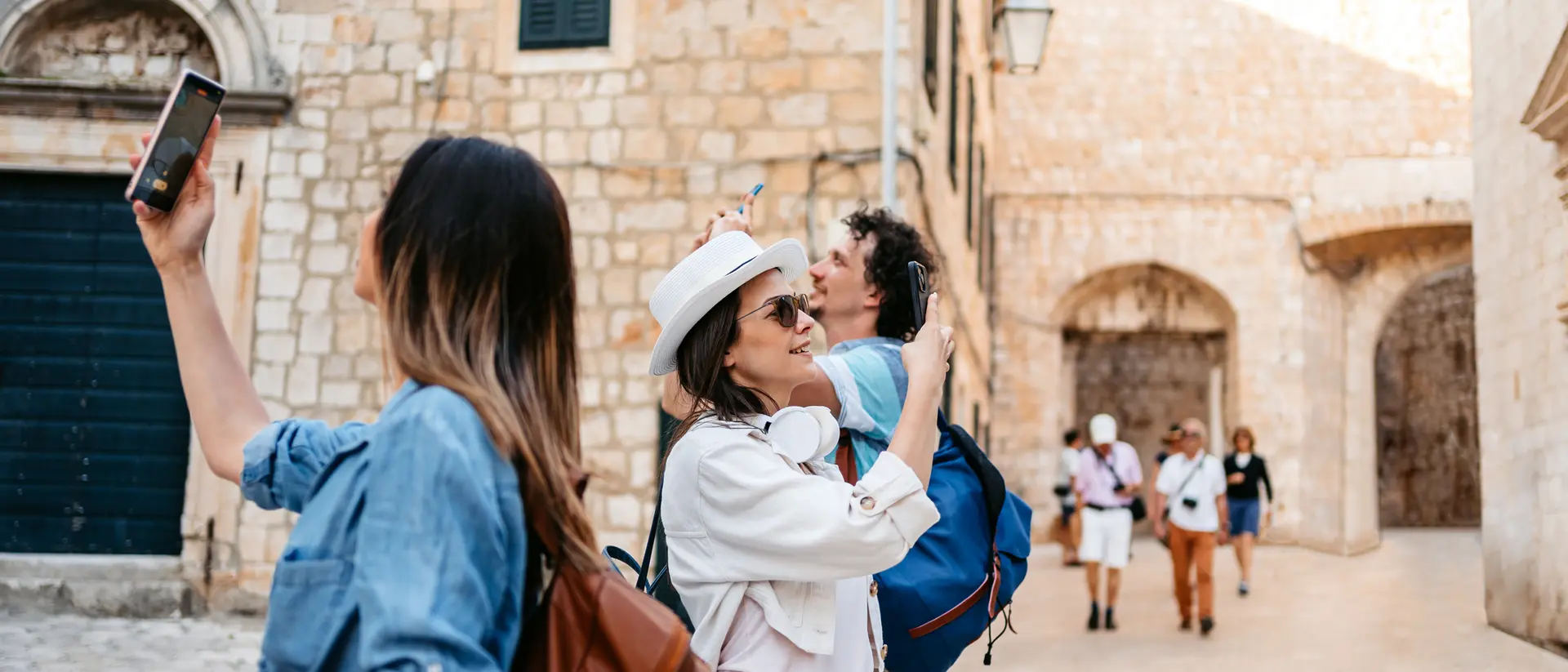 Two women hold phones while taking photos in a picturesque, stone-walled street, with others walking nearby.