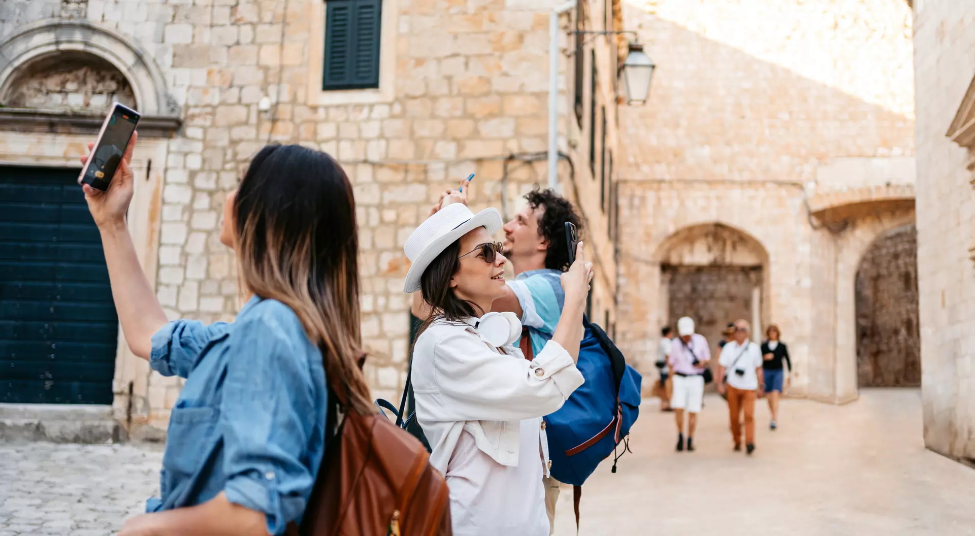 Two women hold phones while taking photos in a picturesque, stone-walled street, with others walking nearby.