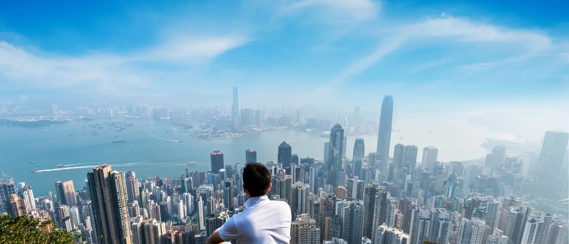 A person sits on a rocky hilltop, overlooking a sprawling city skyline under a clear blue sky.