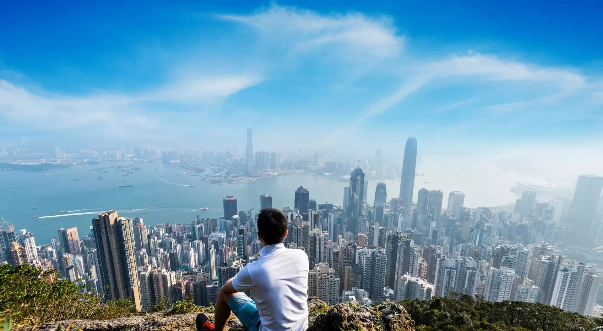 A person sits on a rocky hilltop, overlooking a sprawling city skyline under a clear blue sky.