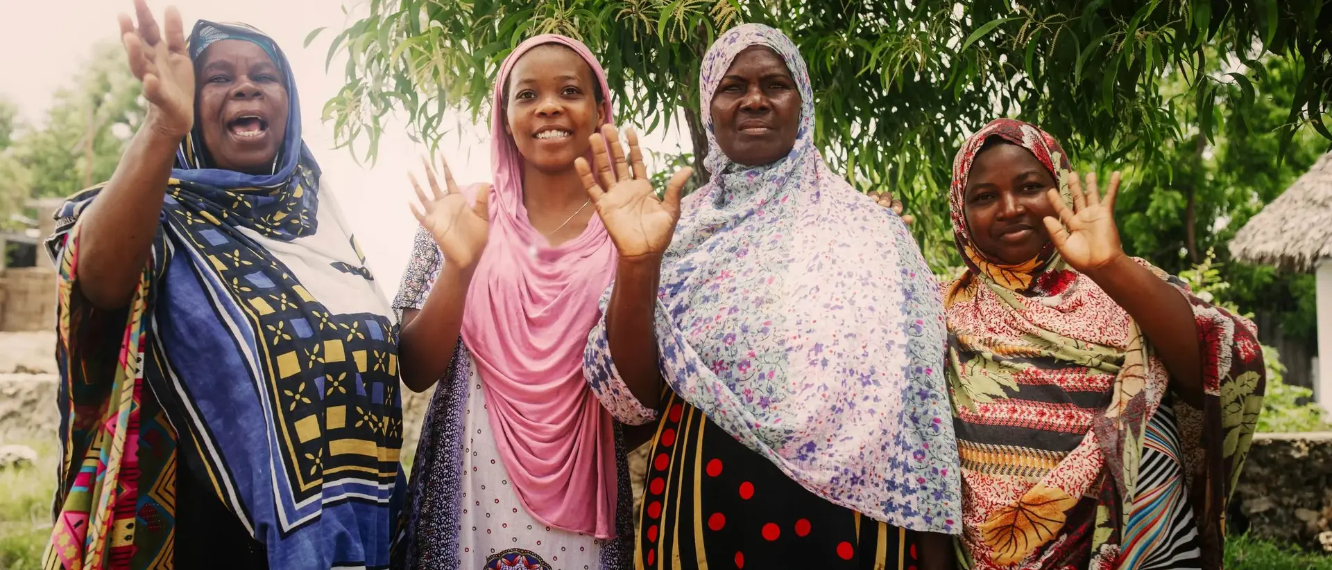 Four women in colorful dresses wave and smile, standing together under a tree in a vibrant outdoor setting.