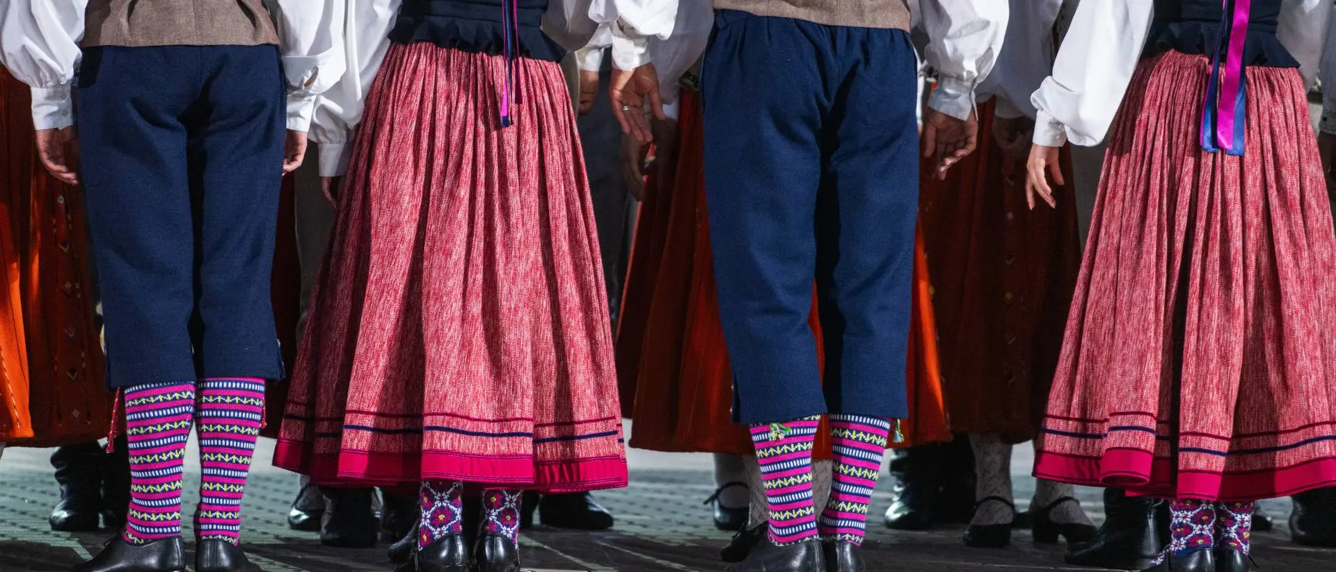 Group of dancers in traditional attire, showcasing colorful skirts and patterned socks from the back.