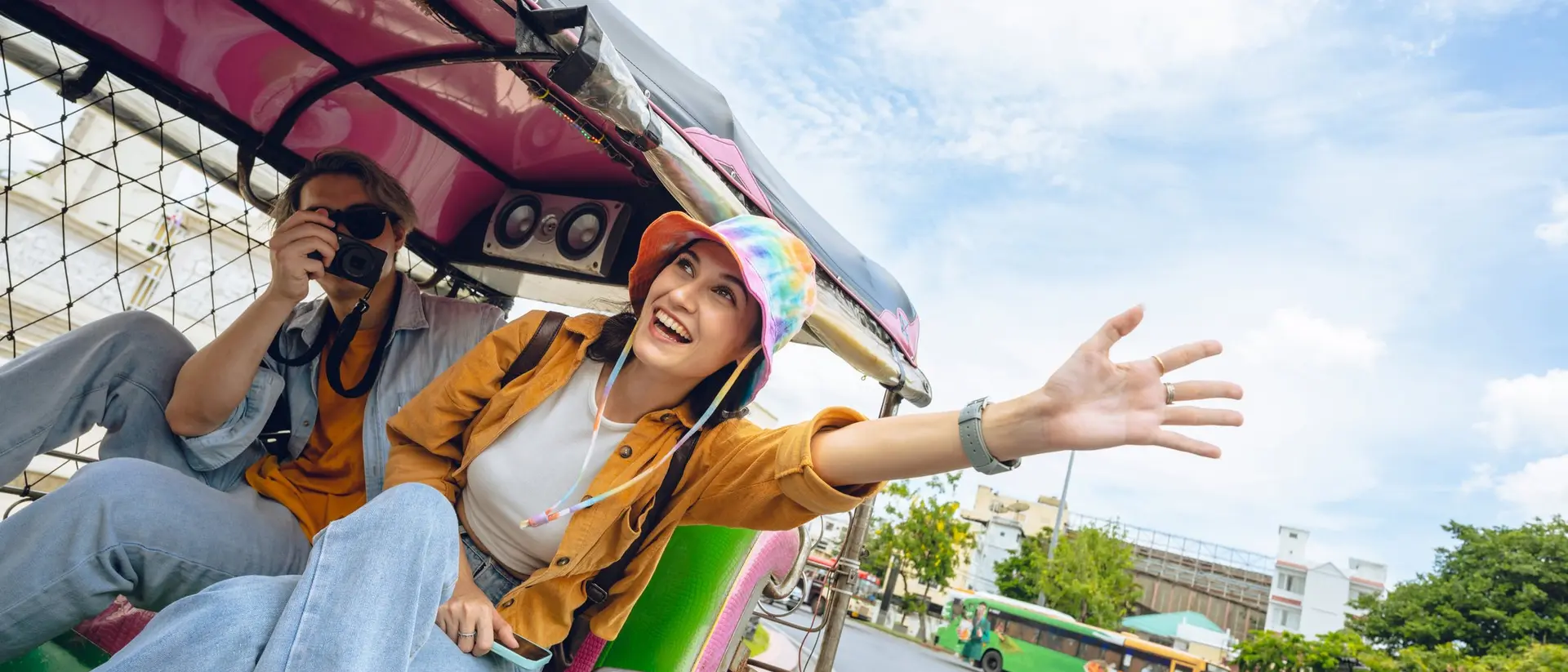 Two friends enjoy a ride in a colorful tuk-tuk, with one waving and smiling while the other takes pictures.