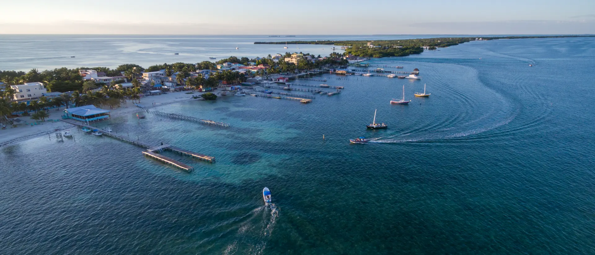 Aerial view of a coastal town with boats navigating clear blue waters and a serene sky in the background.