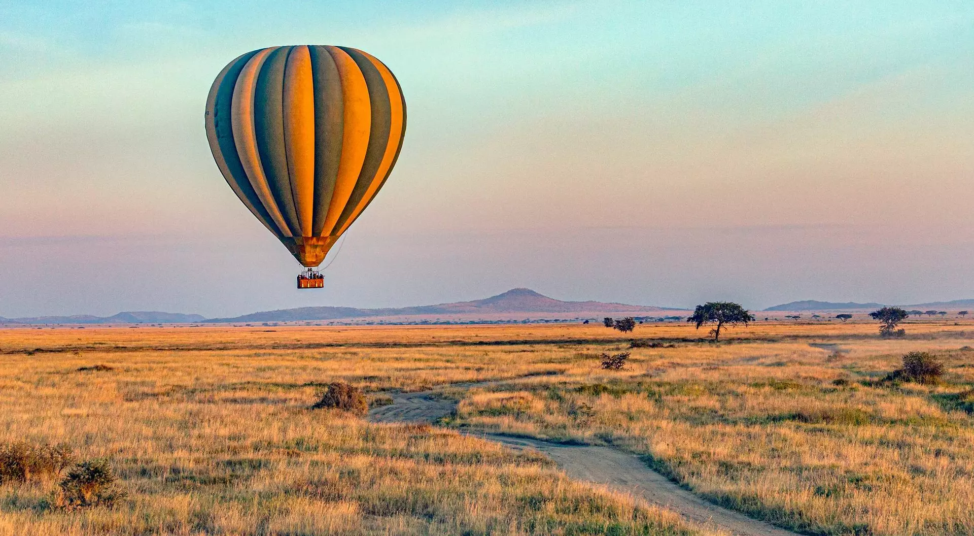 A colorful hot air balloon floats above a vast, grassy landscape under a clear sky. A dirt path winds through the field.