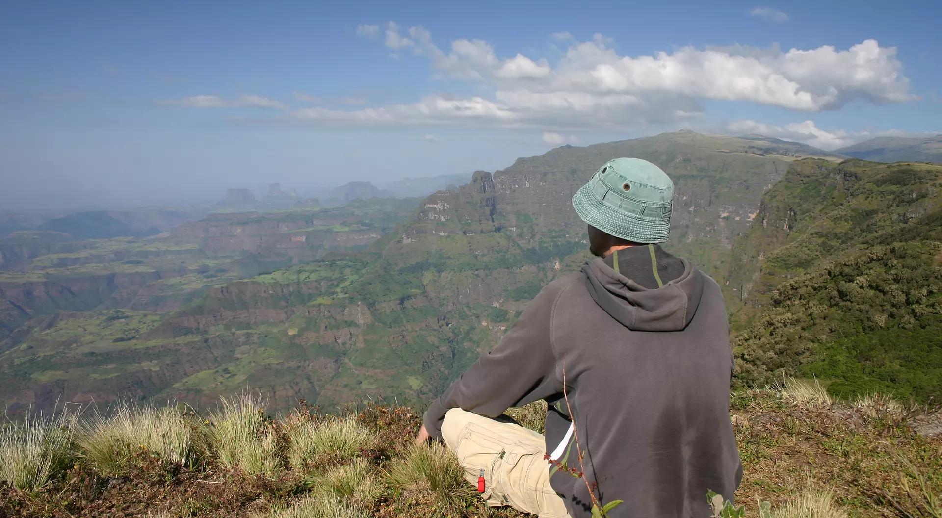 Person in a hat sitting on a mountain overlooking lush, green valleys and distant hills under a clear blue sky.