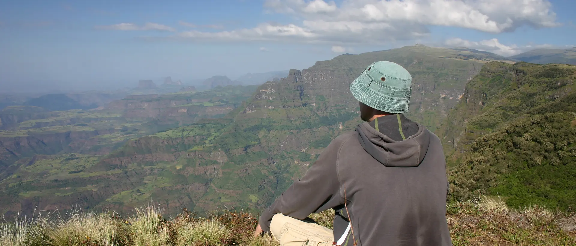 Person in a hat sitting on a mountain overlooking lush, green valleys and distant hills under a clear blue sky.