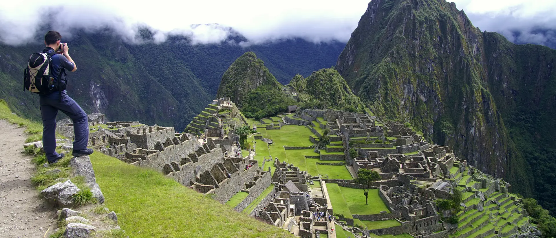 A person stands on a path overlooking the ancient ruins of Machu Picchu, surrounded by green mountains and clouds.