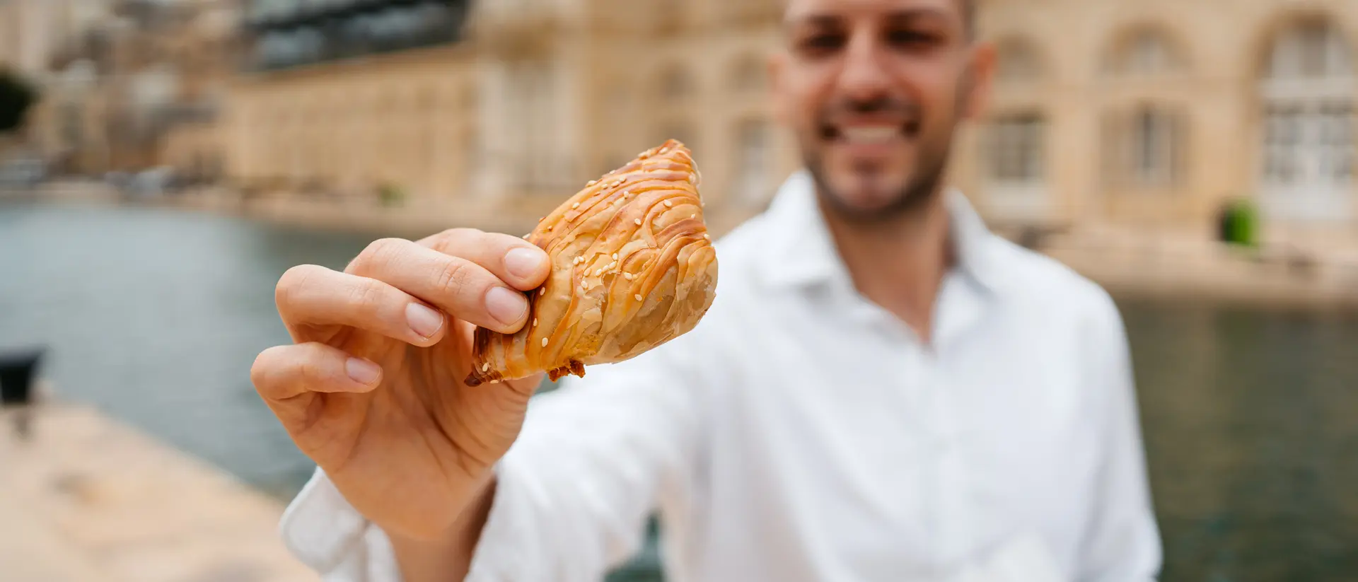 A man in a white shirt holds up a pastry by a calm waterway, smiling in front of a historic building.