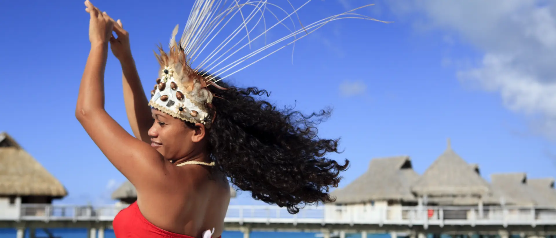 A dancer in a red bikini top and decorative crown sways against a tropical ocean backdrop with thatched huts.
