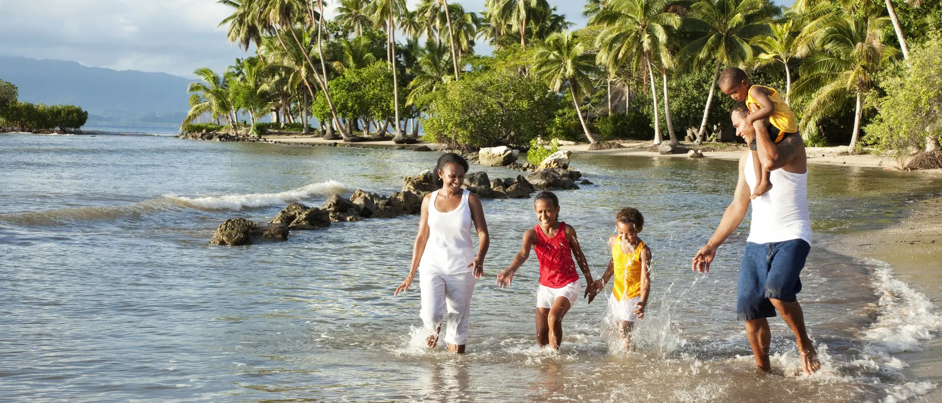 A family plays in shallow water on a beach with palm trees in the background, enjoying a sunny day together.