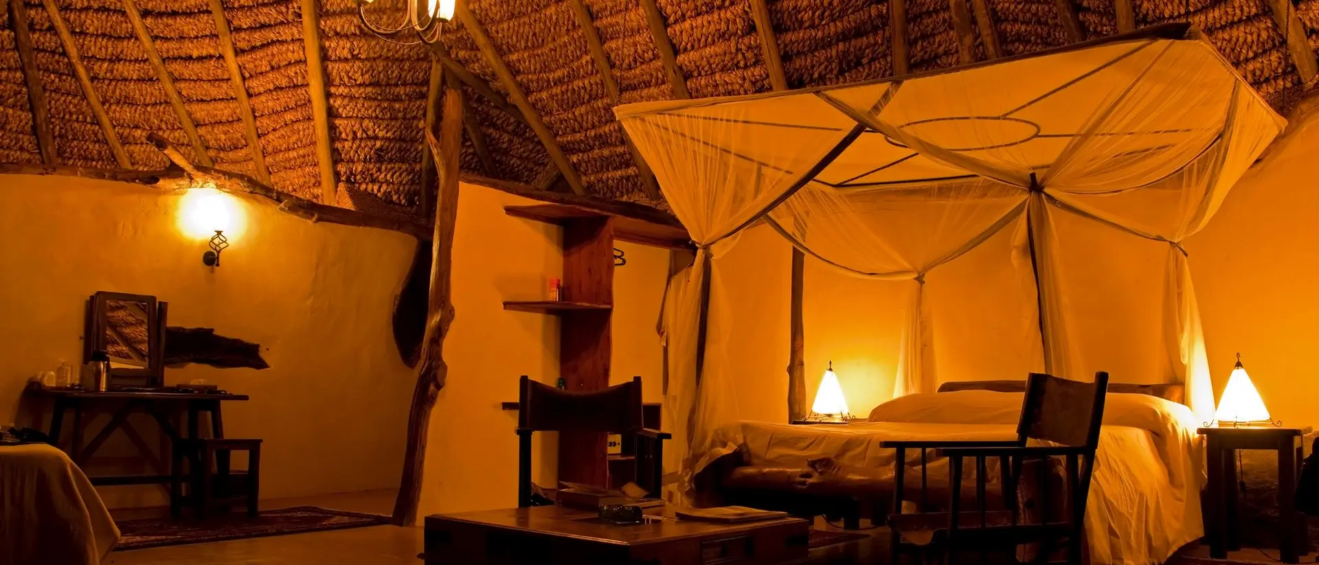 Interior of a cozy, warmly lit room with a thatched roof, featuring a bed with a canopy and rustic furniture.