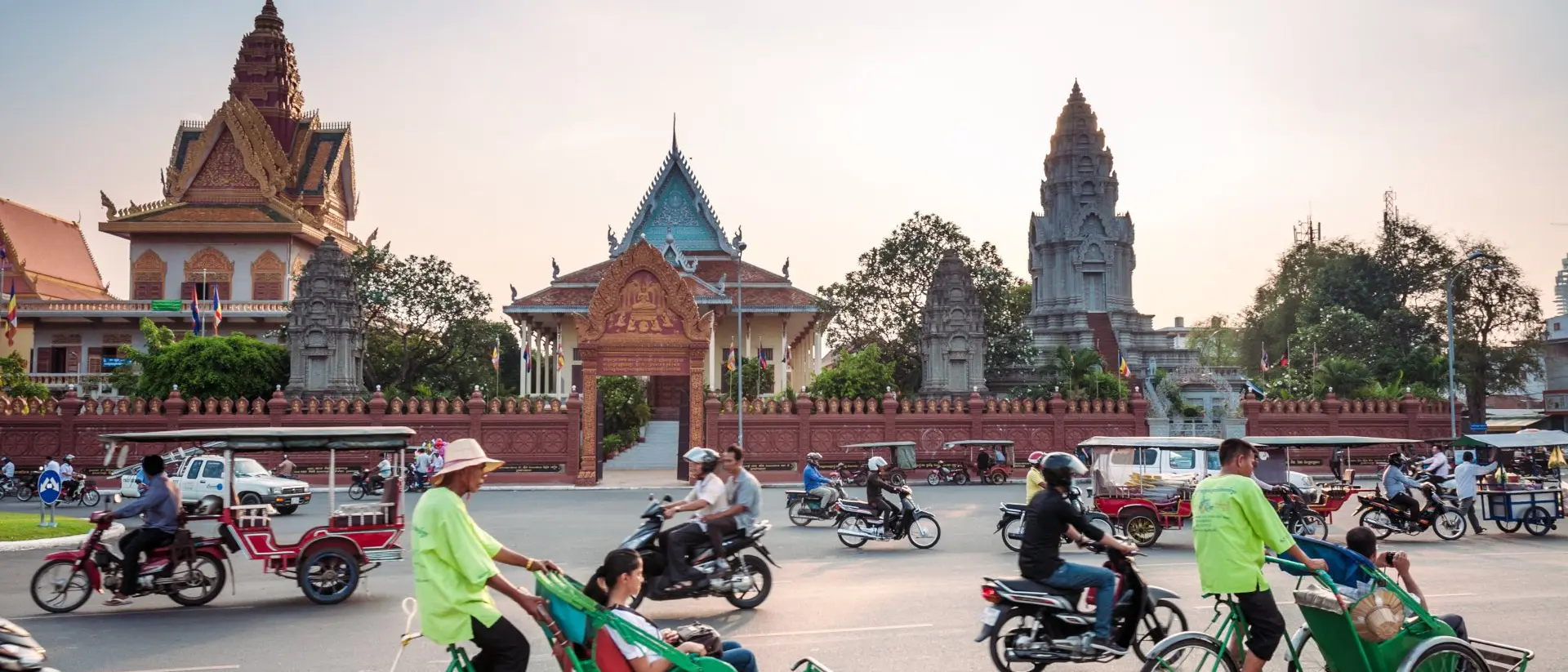 Traffic scene with rickshaws and motorbikes near a colorful temple complex at sunset.