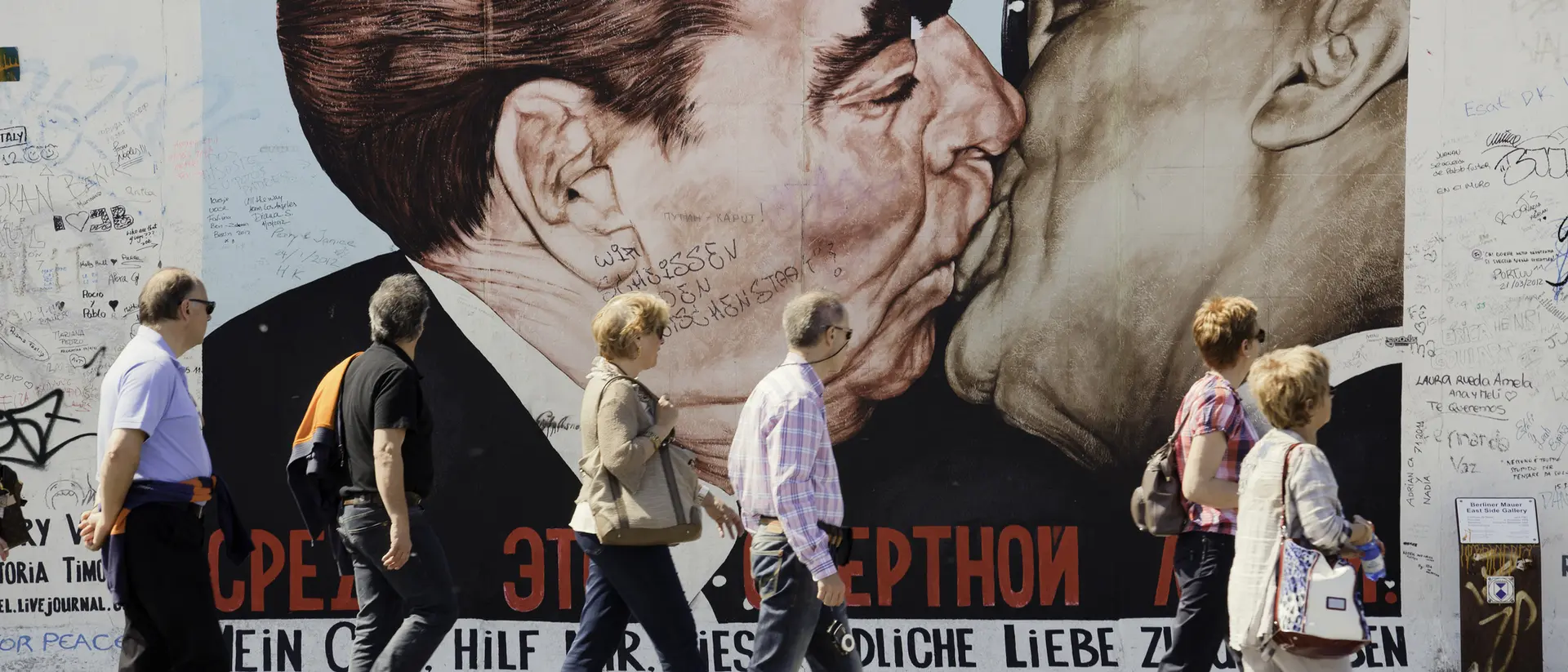 People walk by a large mural of two leaders kissing on a wall, with graffiti surrounding the artwork.