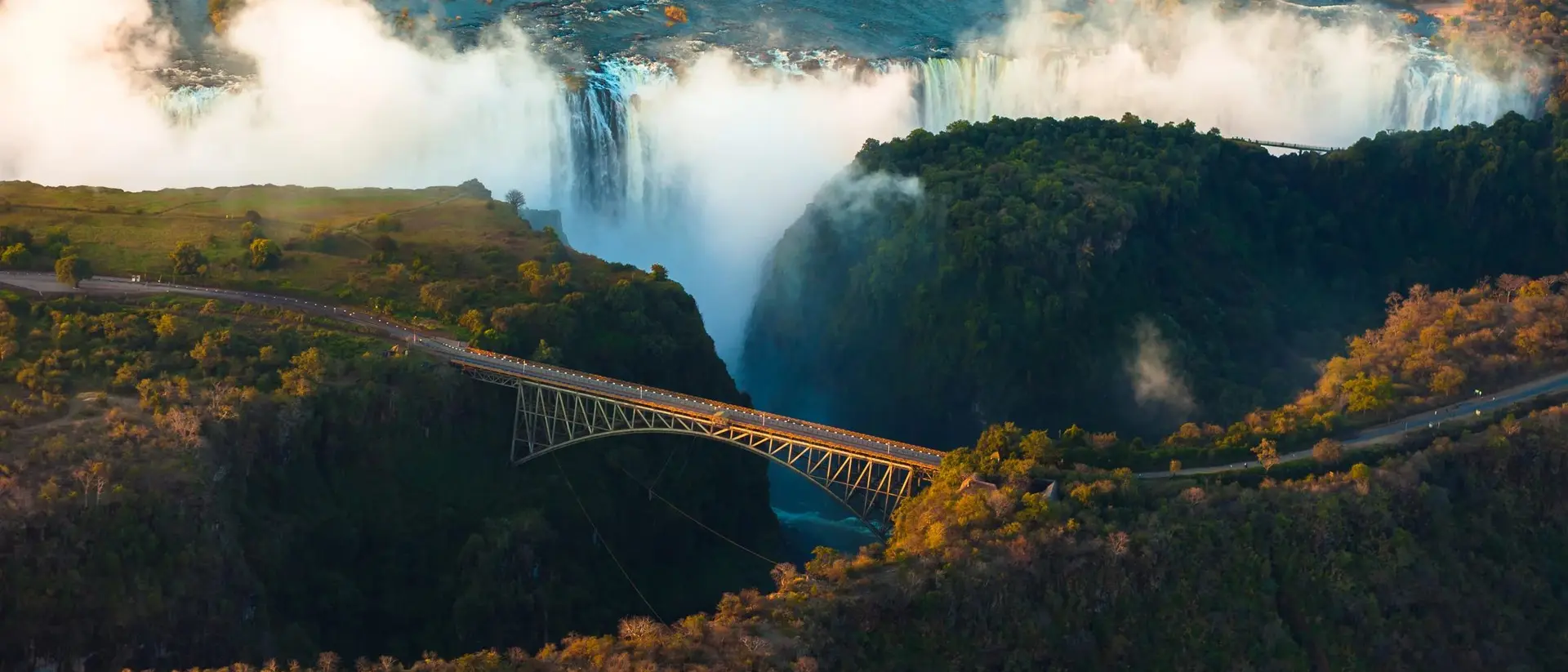 Aerial view of a waterfall with mist, featuring a bridge spanning a canyon surrounded by lush greenery.