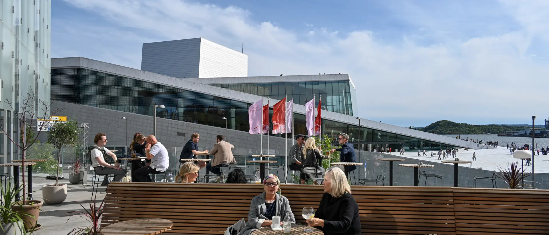 People relax on a sunny terrace outside a modern building, enjoying drinks and conversation. Flags flutter in the background.