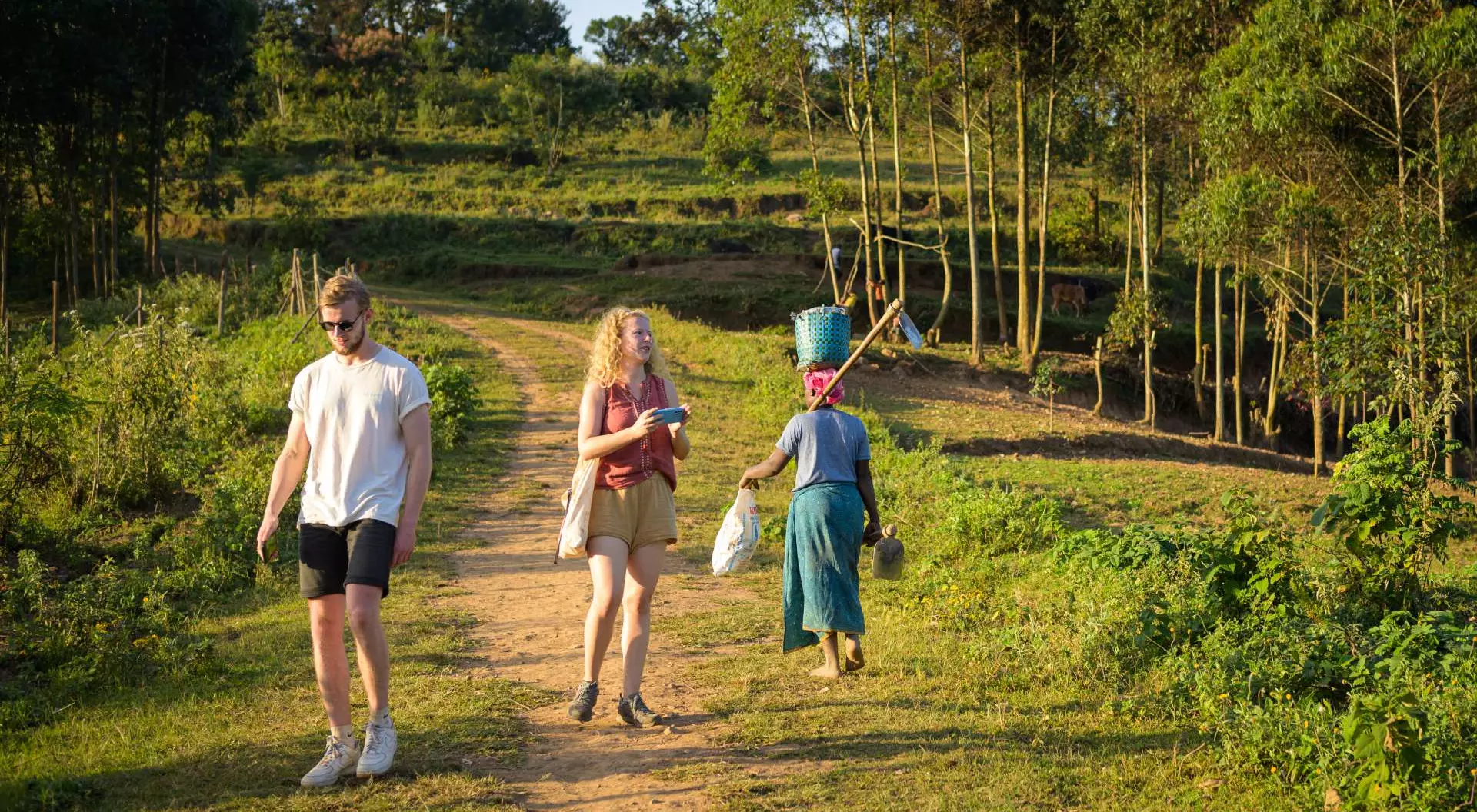 Two people walk on a dirt path in a rural setting, while a woman carries items in a basket nearby. Lush greenery surrounds them.