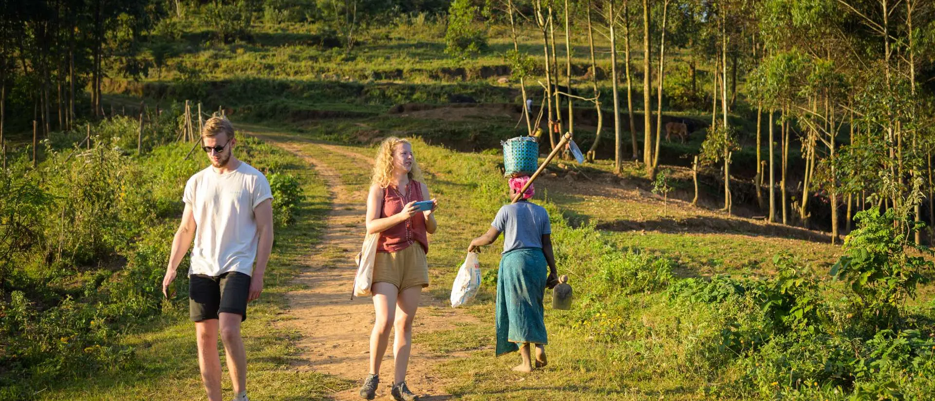 Two people walk on a dirt path in a rural setting, while a woman carries items in a basket nearby. Lush greenery surrounds them.