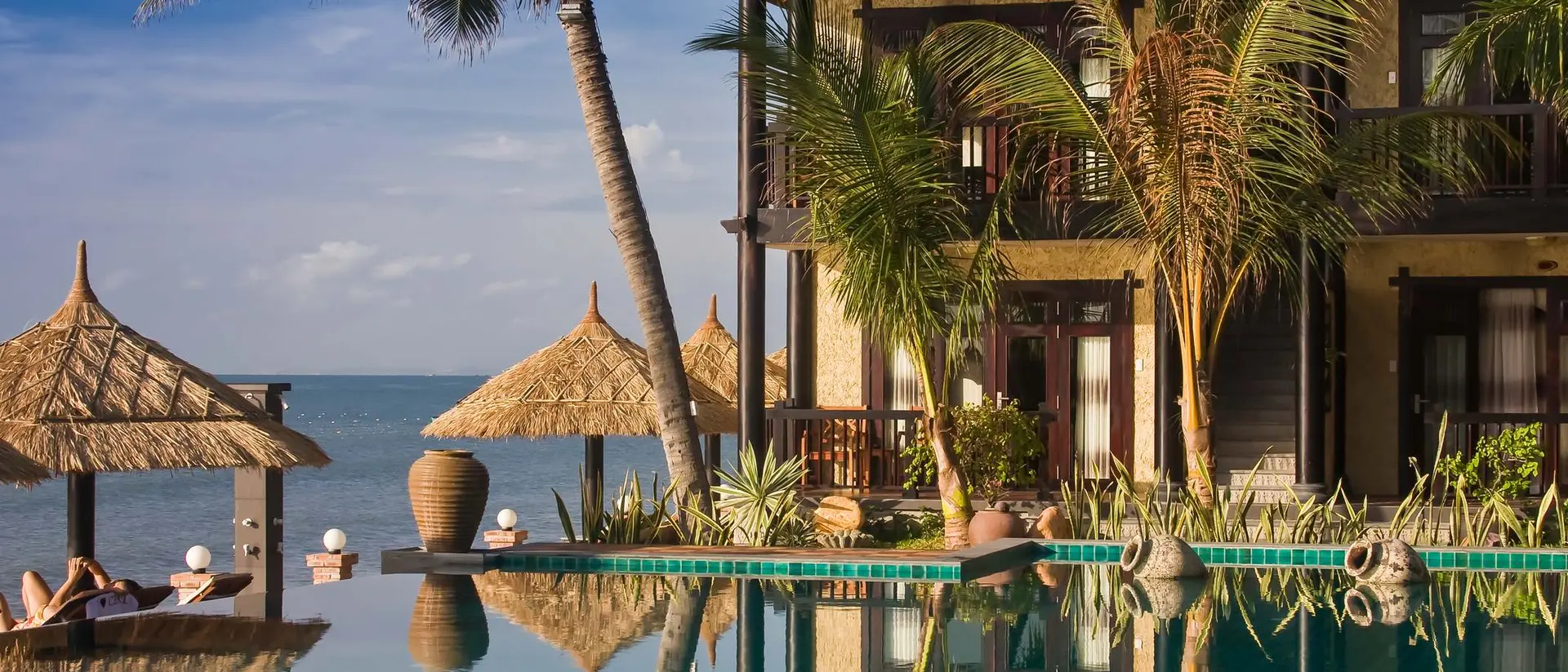 Tropical beachfront villa with palm trees, a pool, and thatched umbrellas overlooking the ocean at sunset.