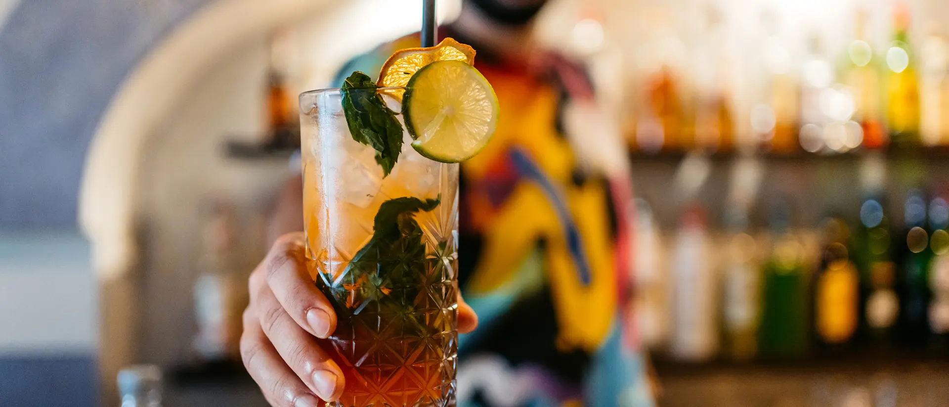 A bartender holding a colorful cocktail garnished with mint and citrus, with shelves of bottles in the background.
