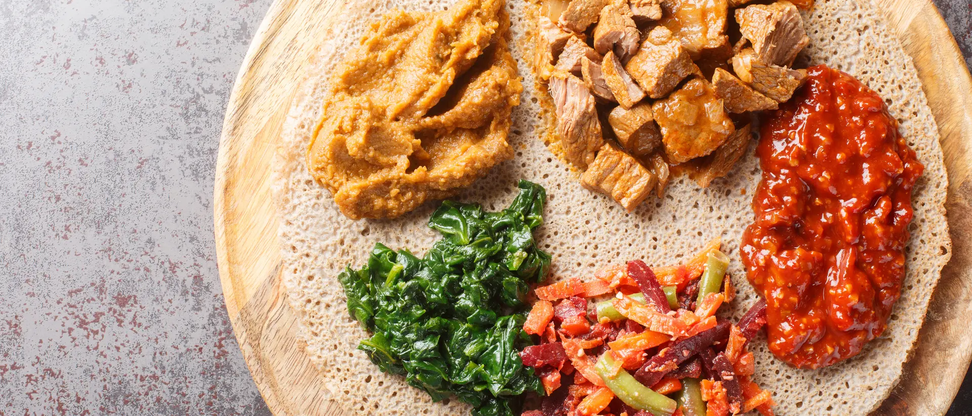 A wooden platter with injera topped with various dishes: spiced meat, lentils, greens, and a colorful vegetable mix.