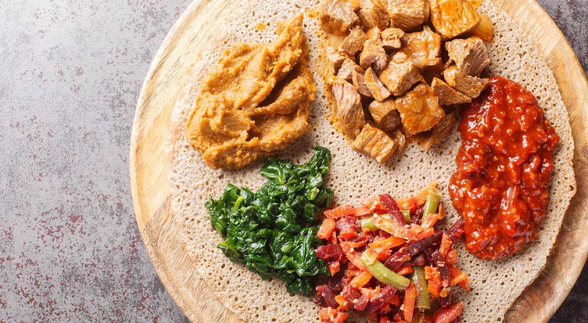 A wooden platter with injera topped with various dishes: spiced meat, lentils, greens, and a colorful vegetable mix.