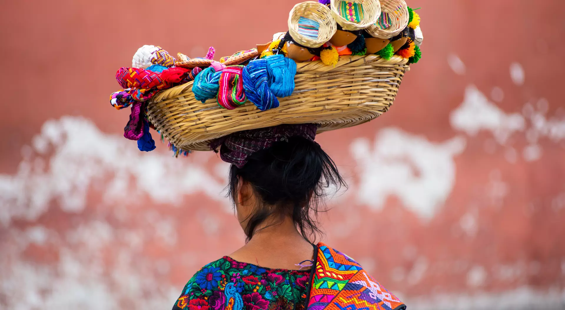 A woman with dark hair carries a colorful woven basket on her head, adorned with crafts and textiles, against a textured wall.