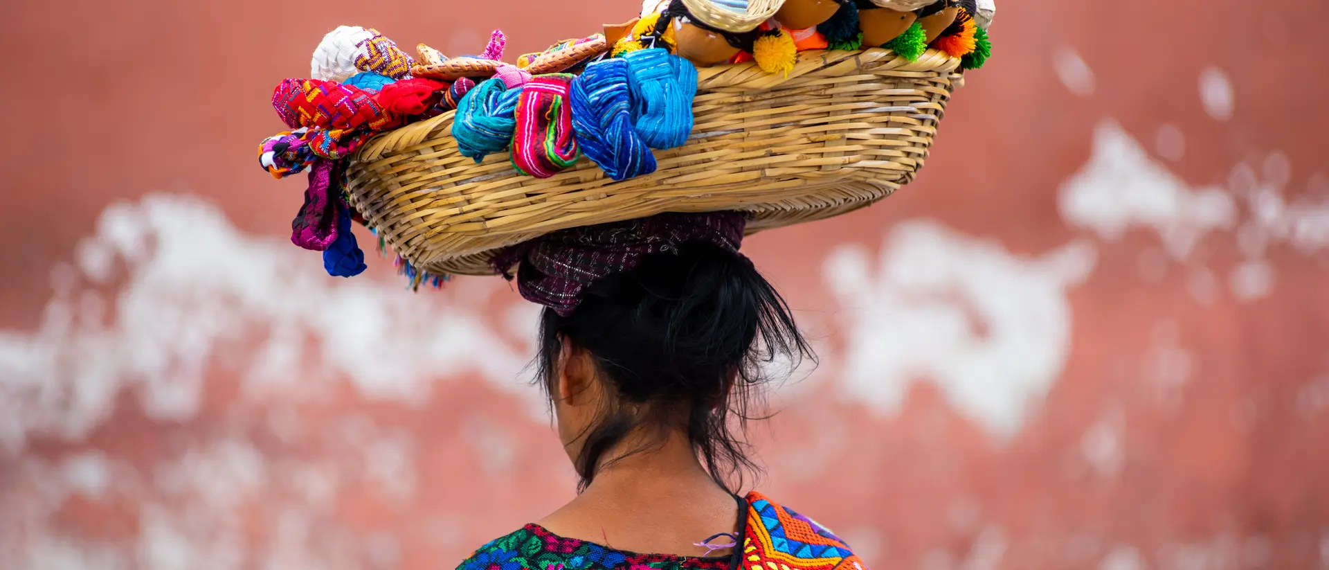 A woman with dark hair carries a colorful woven basket on her head, adorned with crafts and textiles, against a textured wall.