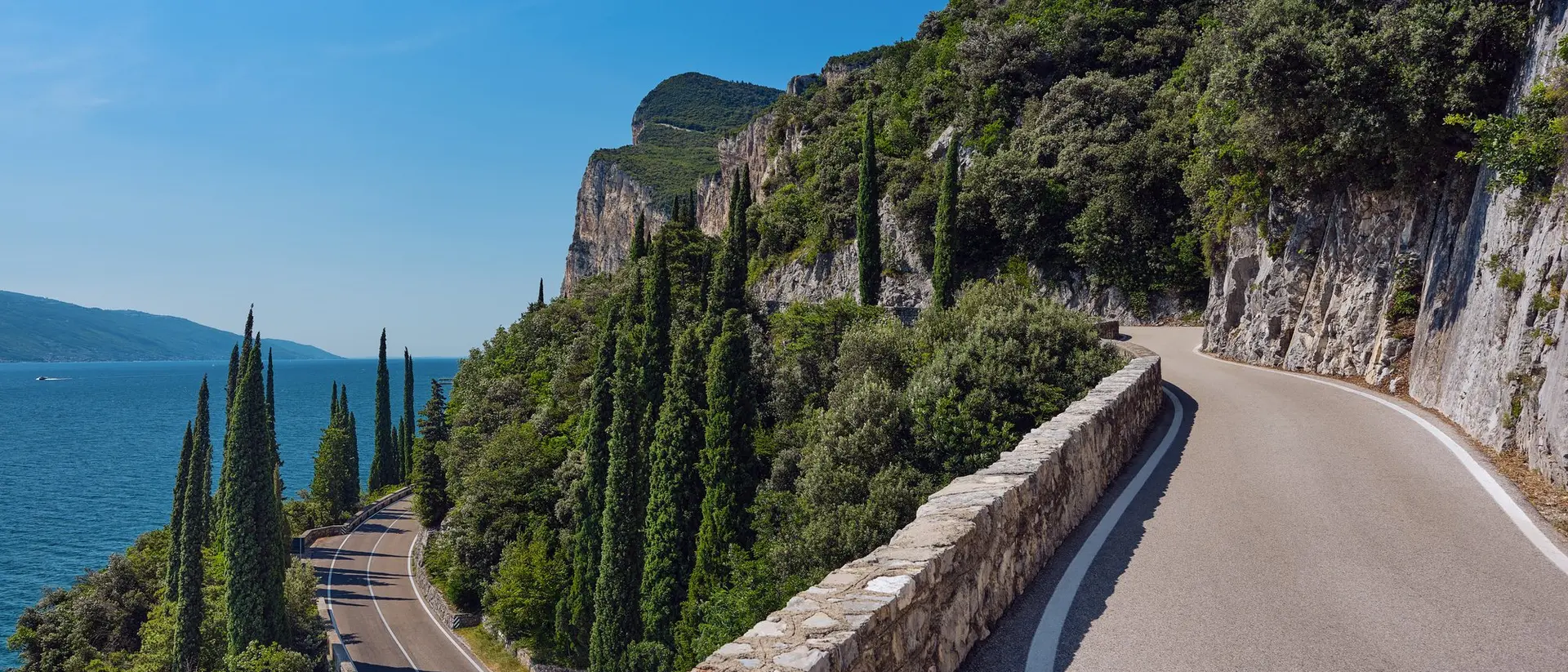 Scenic winding road along a cliffside with lush greenery and a view of the lake under a clear blue sky.