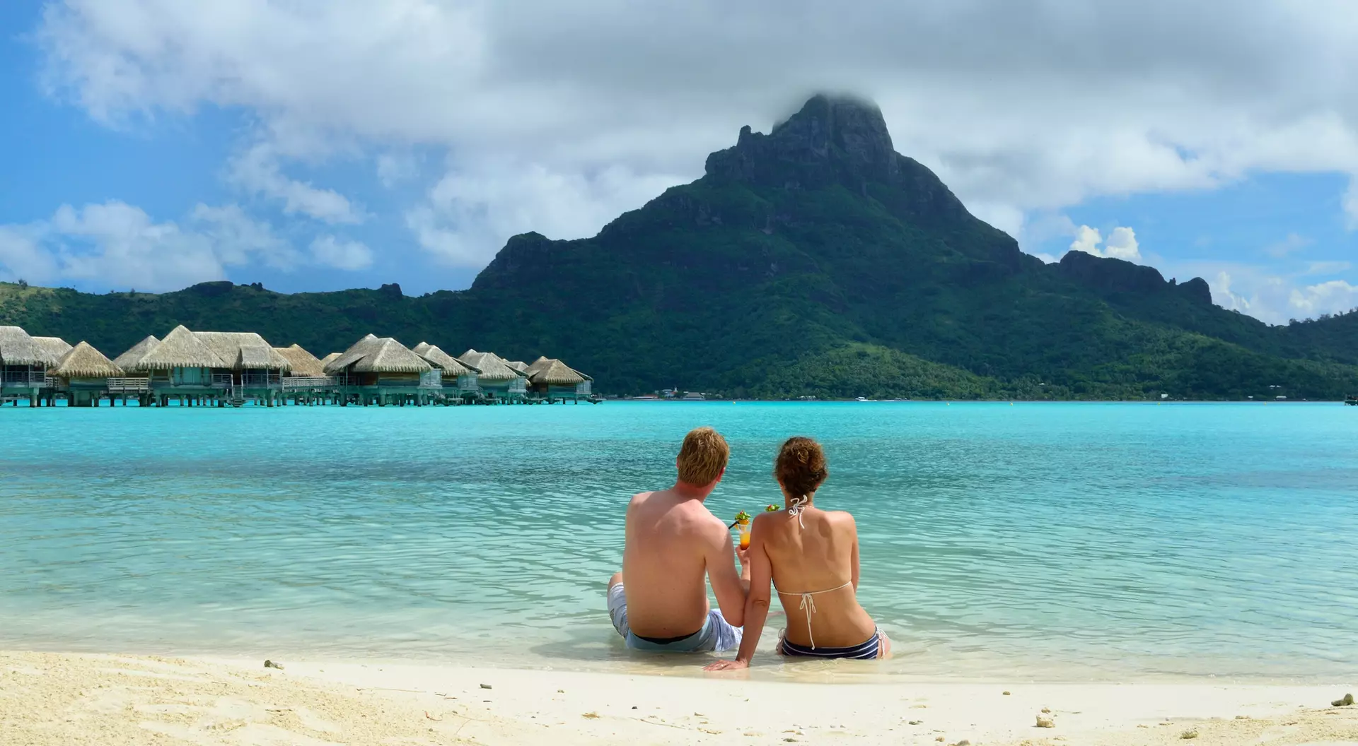 A couple sits on a beach, facing a mountain and turquoise lagoon, with huts in the background and a cloudy sky above.