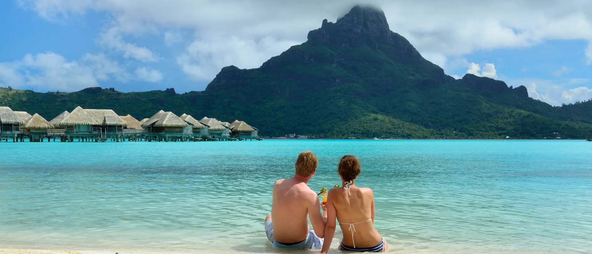 A couple sits on a beach, facing a mountain and turquoise lagoon, with huts in the background and a cloudy sky above.