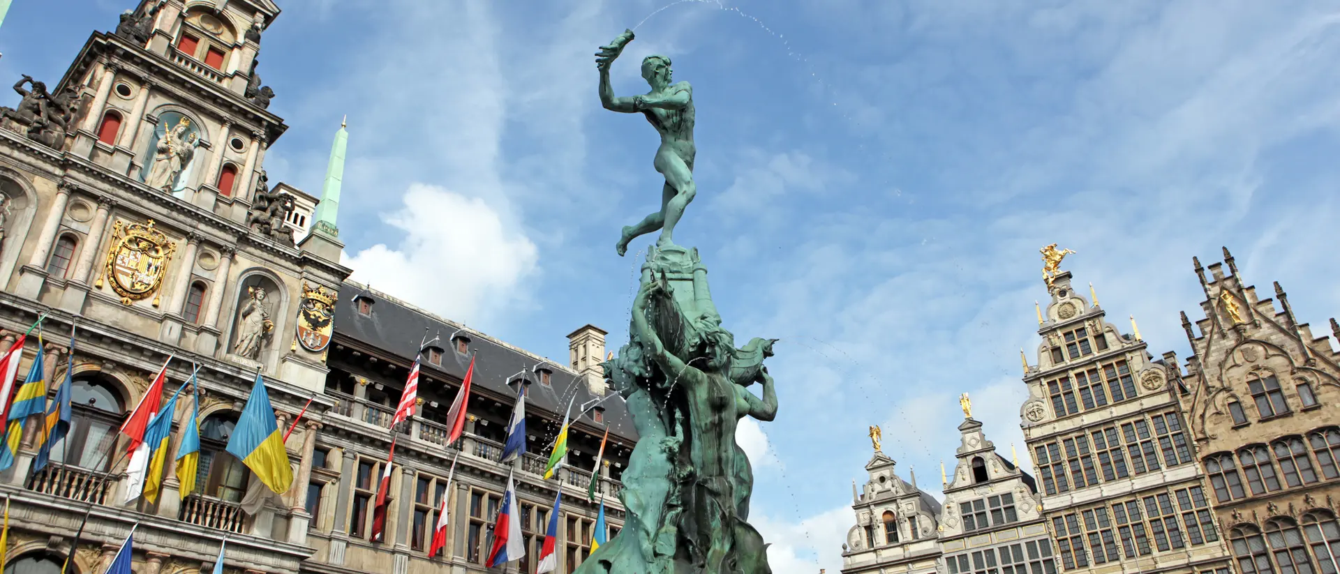 A bronze statue of a man atop a pedestal, surrounded by historic buildings and colorful flags under a blue sky.
