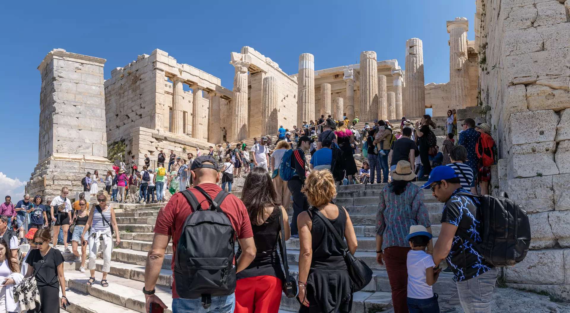 Crowd of tourists ascending steps towards ancient ruins of the Acropolis under a clear blue sky.