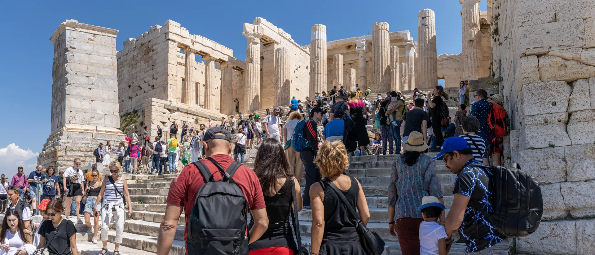 Crowd of tourists ascending steps towards ancient ruins of the Acropolis under a clear blue sky.