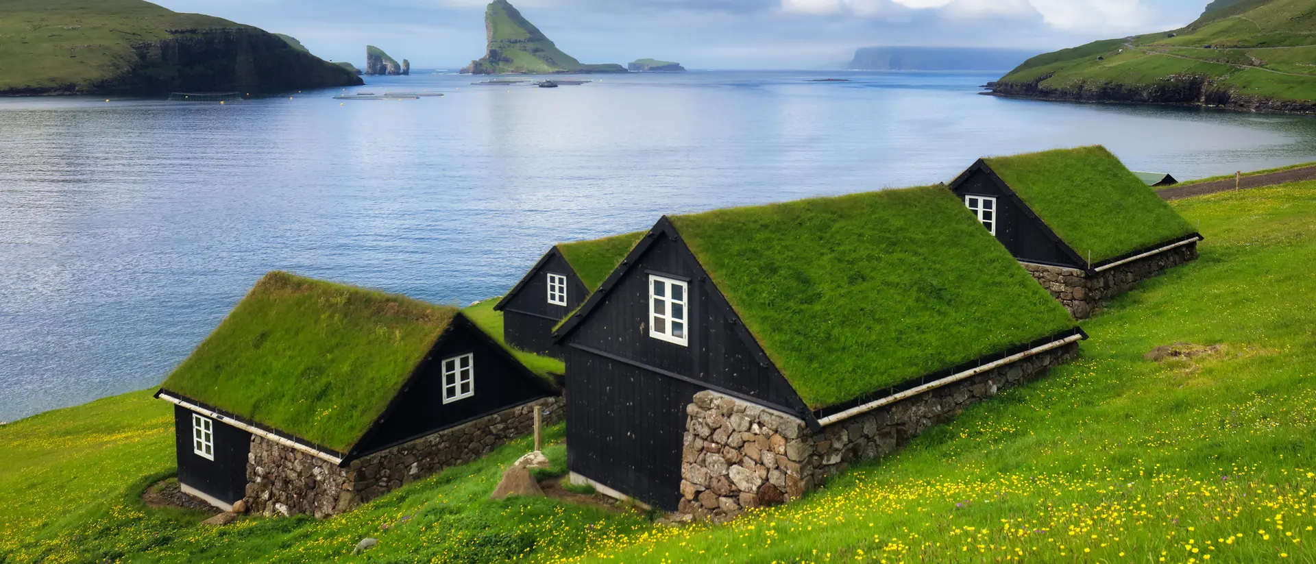 Black cottages with grass roofs on a green hillside overlooking a calm sea and distant rocky islands under a cloudy sky.