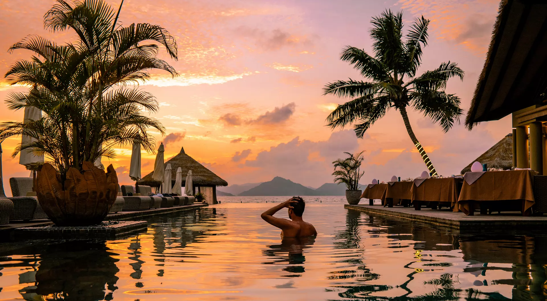 A person relaxes in a pool at sunset, surrounded by palm trees and an idyllic tropical landscape.