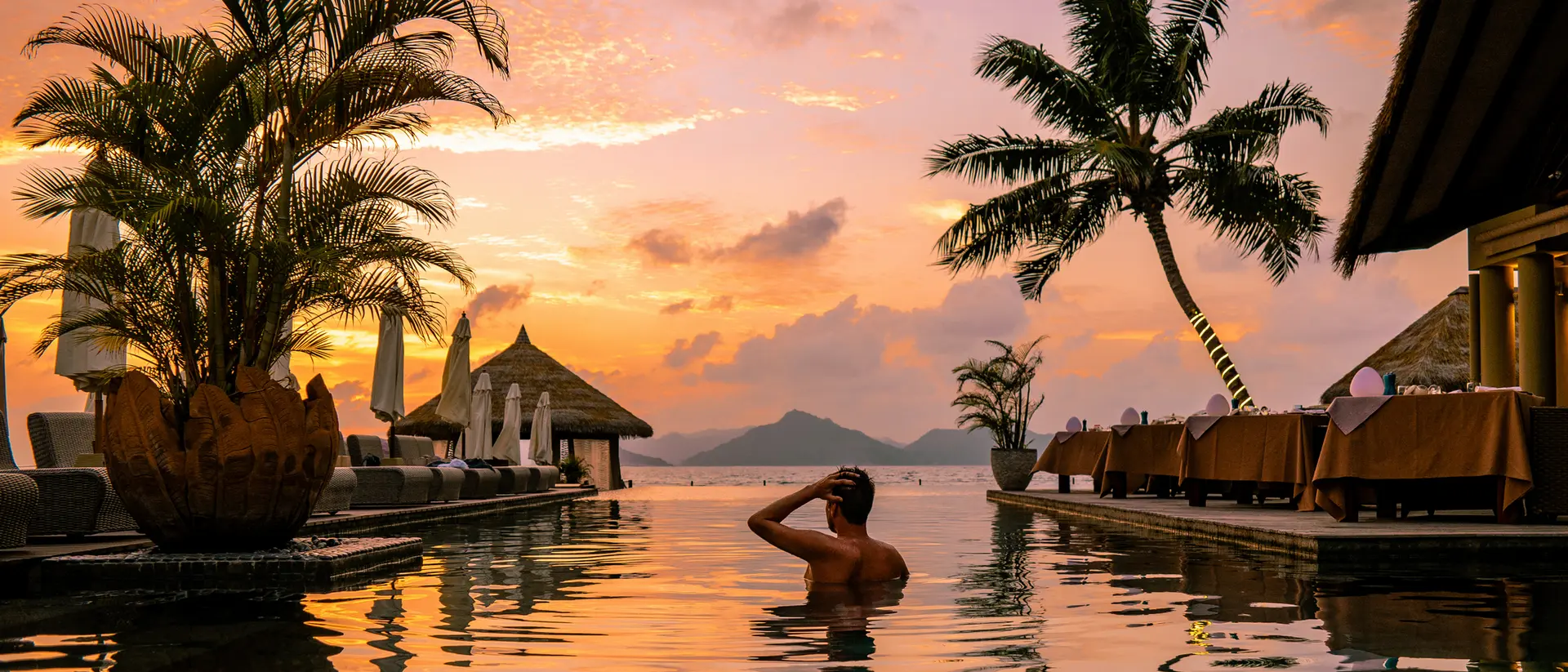 A person relaxes in a pool at sunset, surrounded by palm trees and an idyllic tropical landscape.
