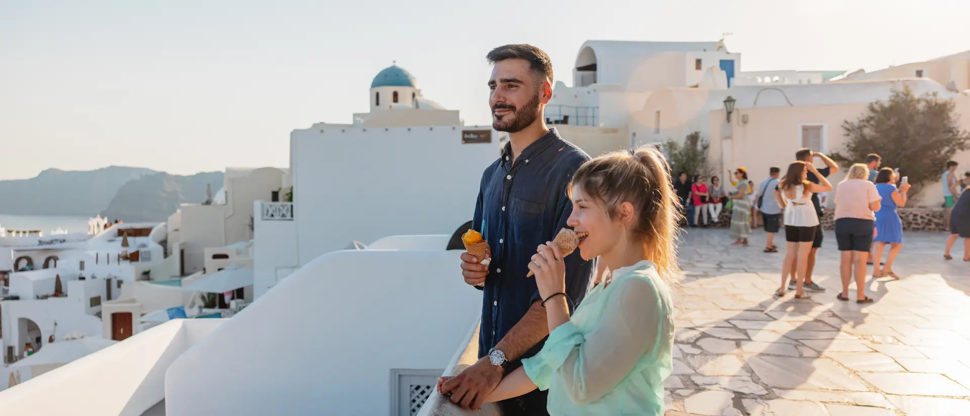 A man and a woman enjoy ice cream on a balcony, overlooking white buildings and people in a sunny coastal landscape.