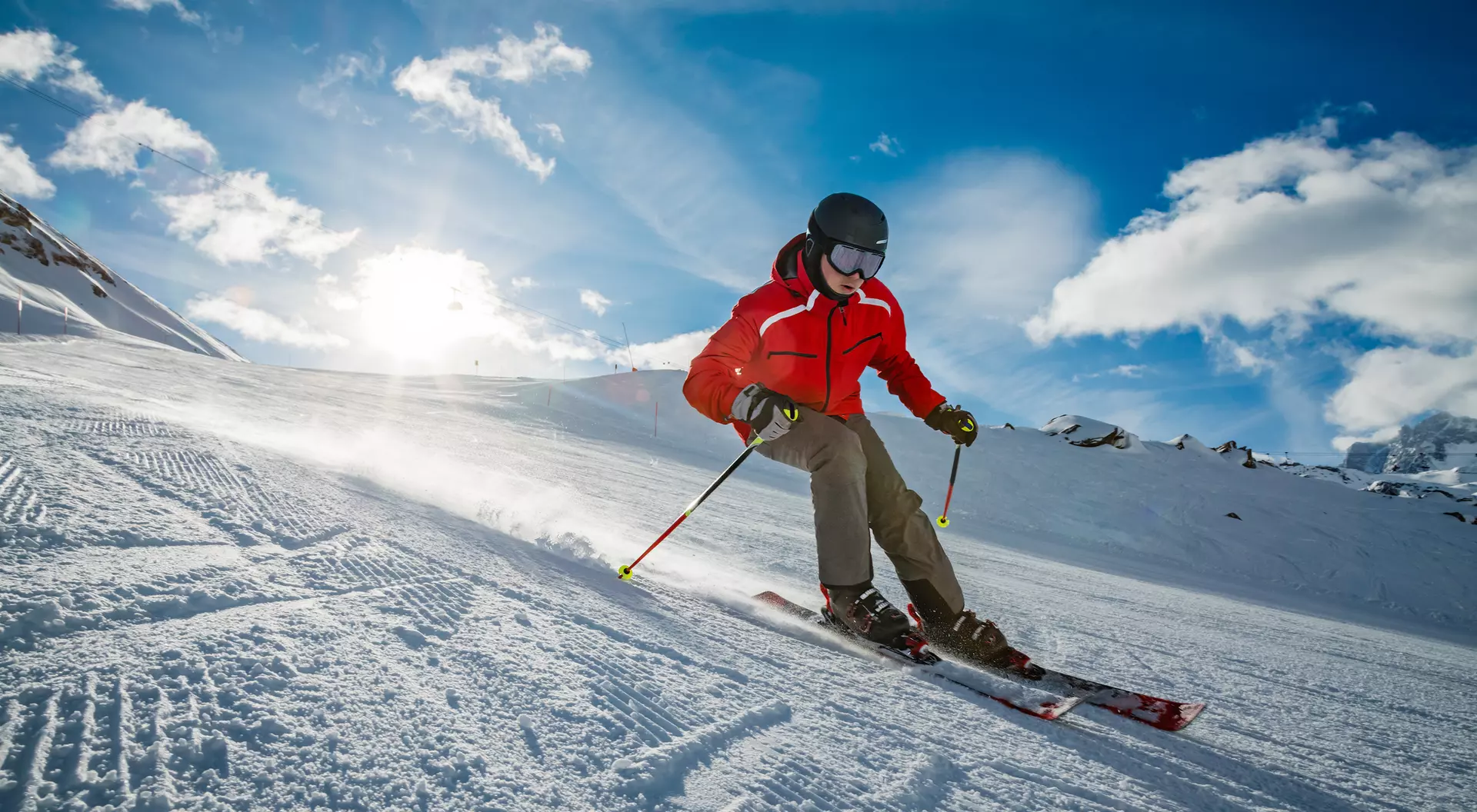 A skier in a red jacket glides downhill on snowy terrain under a bright blue sky with clouds.