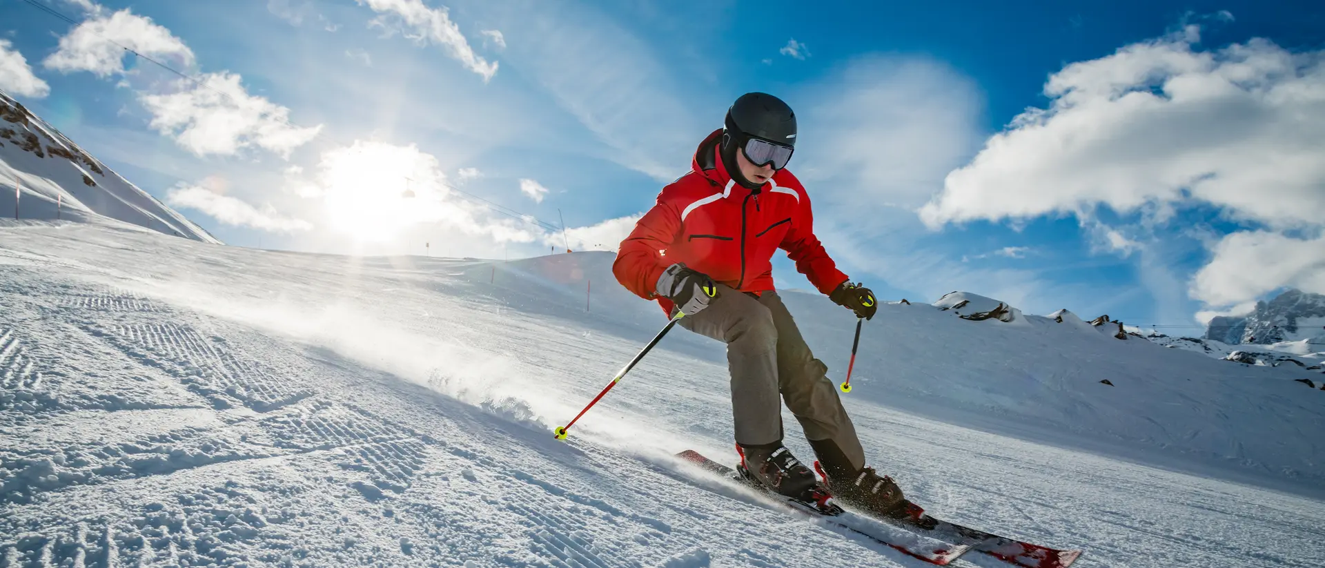 A skier in a red jacket glides downhill on snowy terrain under a bright blue sky with clouds.