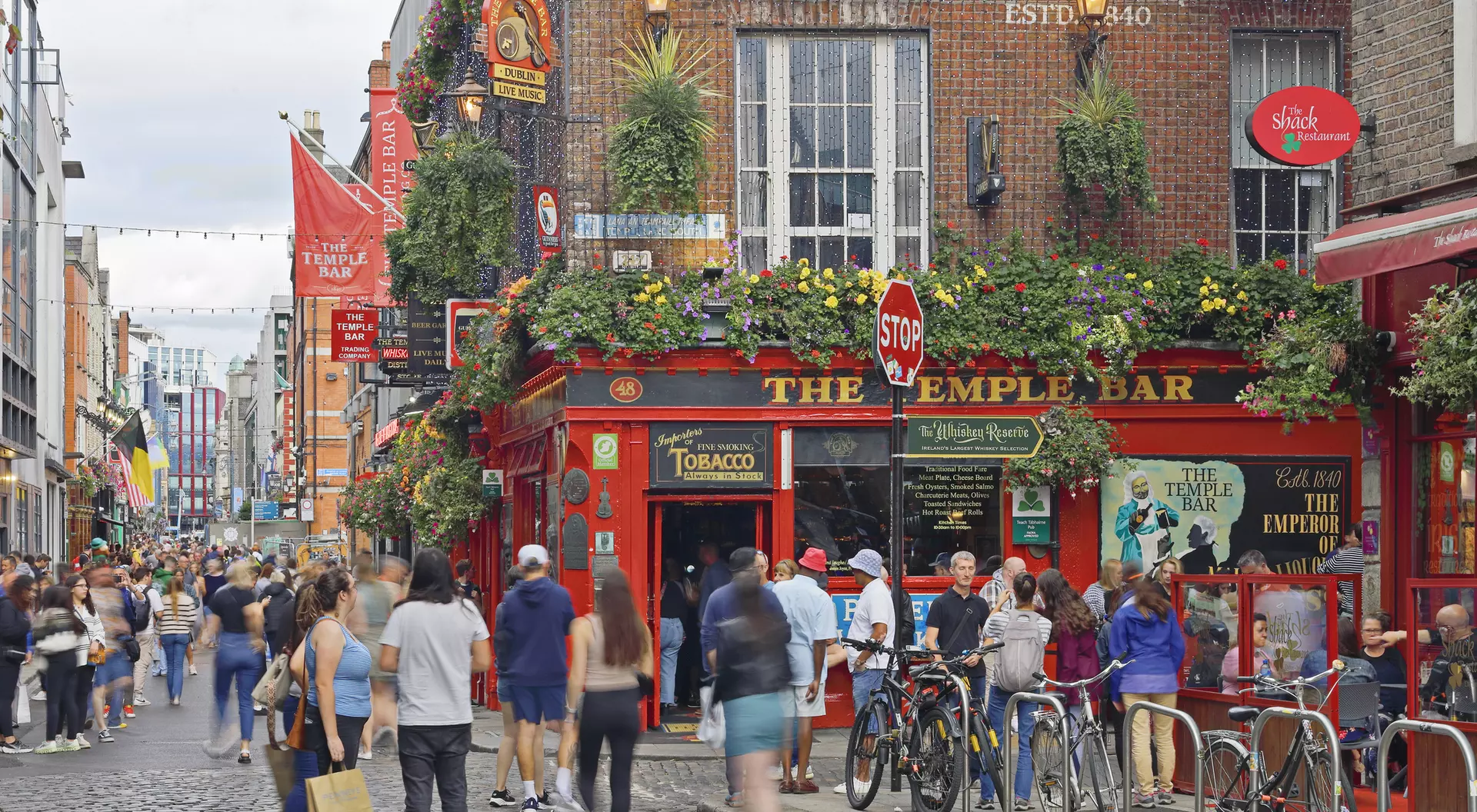 Busy street scene with people walking by the Temple Bar pub, adorned with greenery and colorful banners.